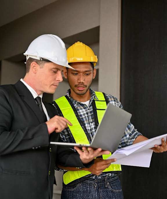 Two construction workers, one in a white hard hat and the other in a yellow hard hat, are reviewing documents together.