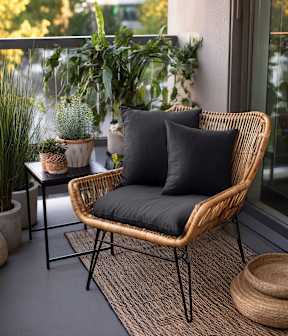 A wicker chair with a black cushion sits on a grey carpet in front of a window with a view of greenery outside.