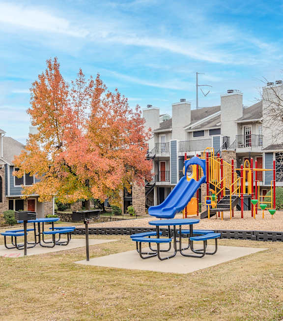 A playground with a blue slide and picnic tables in front of apartment buildings.