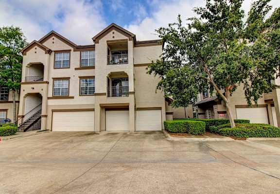 front view of an apartment building with garages and trees at Stone Creek at Old Farm Apartments, Texas