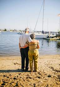 An elderly couple stand on the beach looking out at the water.