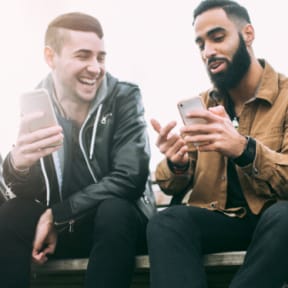 two men sitting on a bench looking at their cell phones