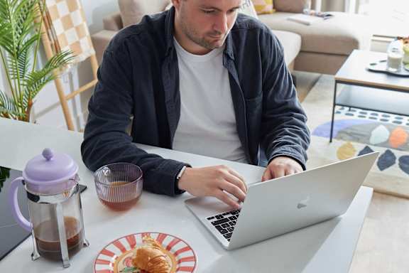 A man is working on his laptop at a table with a purple kettle and a plate of food.