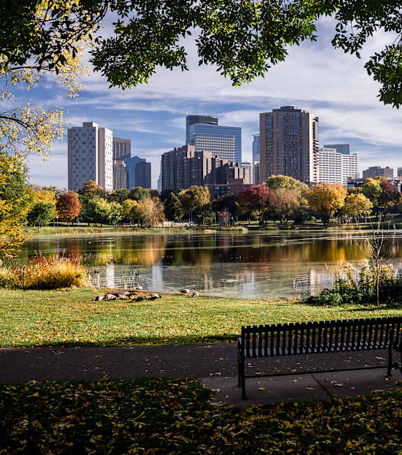 Two black benches are in the foreground of a city park with a lake and trees.