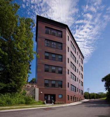 A tall red brick building with many windows sits next to a tree and a road.