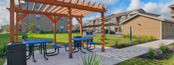 A wooden pergola with blue picnic tables and a trash can is in the foreground of a residential area.