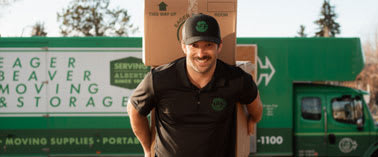 A man in a black shirt and cap is carrying a box while standing in front of a green moving truck.