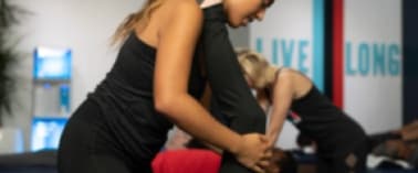 A woman in a black top and leggings is assisting another woman in a yoga pose.