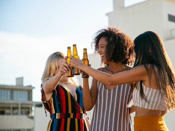 Three women toasting with beer bottles on a rooftop.