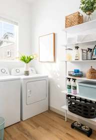 A white laundry room with a washer and dryer, a shelf with cleaning supplies, and a basket plant.