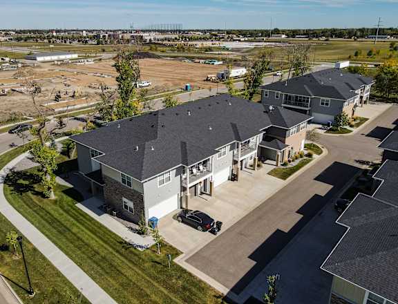 A large grey house with a black roof is surrounded by a grassy area and other houses.