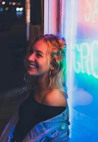 A woman with blonde hair is standing in front of a neon sign.