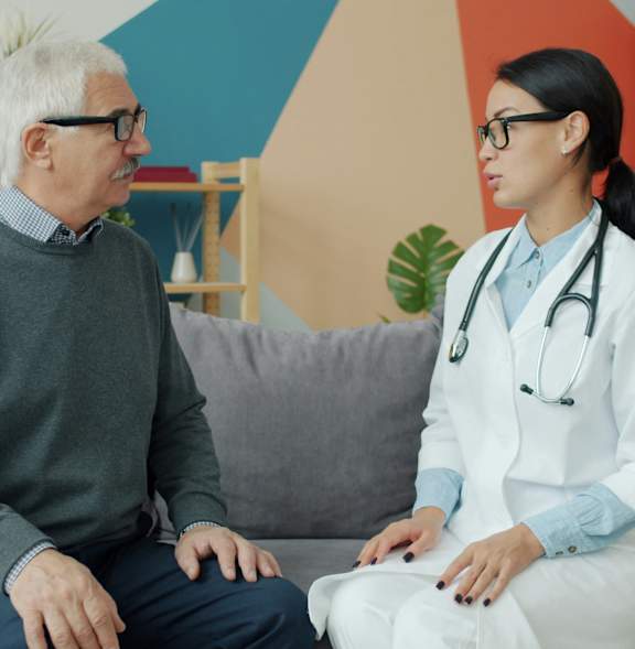 A doctor in a white coat is having a conversation with a patient on a grey couch.