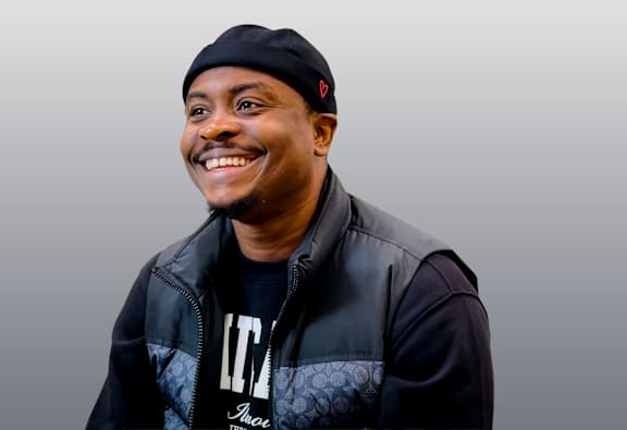 A man wearing a black beanie and a black and grey jacket smiles at the camera.