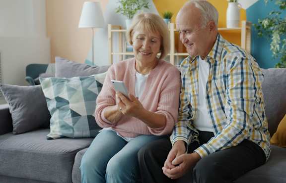An elderly couple is sitting on a grey couch, with the woman holding a smartphone.