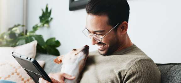A man is sitting on a couch with a dog, using a tablet.