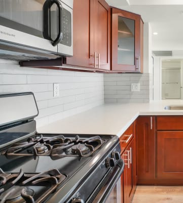 Apartment kitchen with stainless steel appliances, white tile back splash, white counter tops, and cherry wood cabinets.