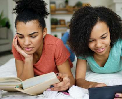 Two people reading a book and using a tablet on a bed.