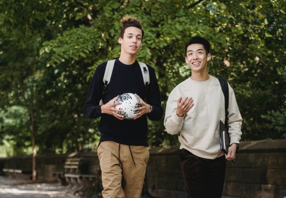 Two men wearing backpacks, one is holding a soccer ball, the other is holding paperwork while walking outdoors down a lush path.