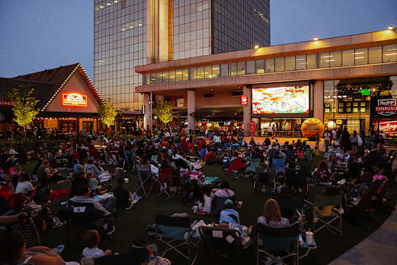 A large crowd of people are sitting on the grass in front of a building with a lit up sign.