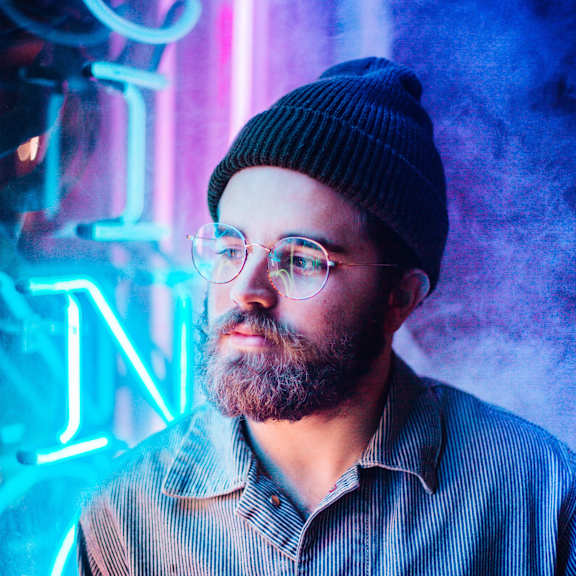 A man with a beard and glasses wearing a beanie and a striped shirt stands in front of a neon sign.
