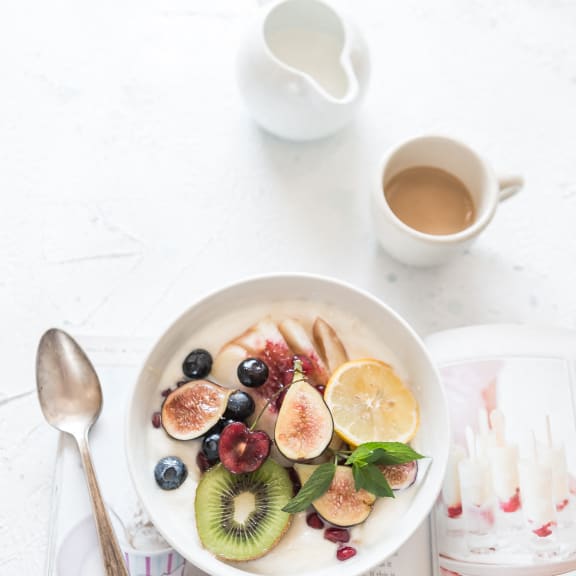 a bowl of fruit on a table with a book and a cup of coffee