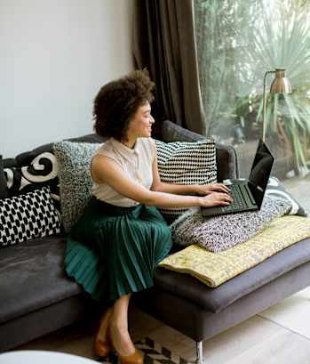 A woman in a green skirt is sitting on a grey couch using a laptop.