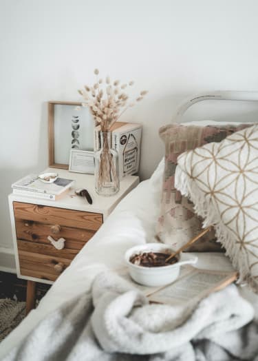 A cozy bedroom scene with a bed, a nightstand with a book, a vase with dried flowers, and a pillow with a geometric pattern at Bradford Gwinnett Townhomes in Norcross, GA