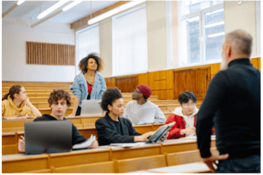 A group of students are sitting in a lecture hall with a teacher standing at the front.