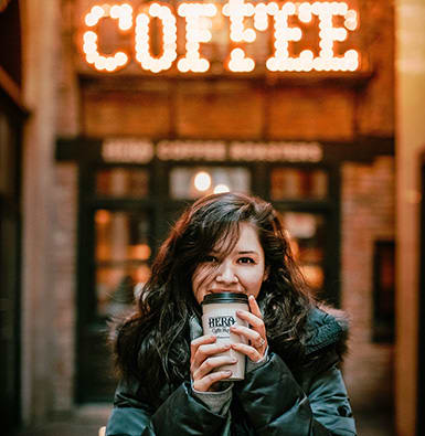 A women in a down jacket clutching a coffee cup in front of a sign saying coffee.