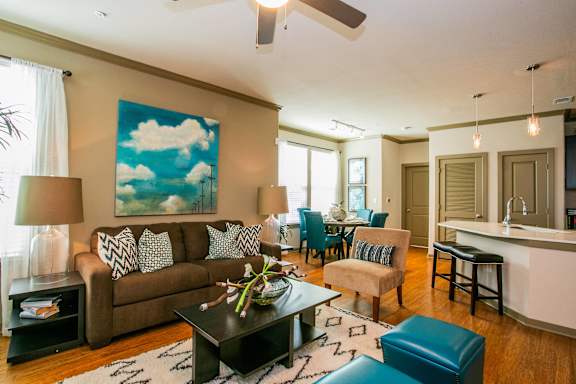 A living room with a brown couch and a painting of a blue sky with clouds on the wall.