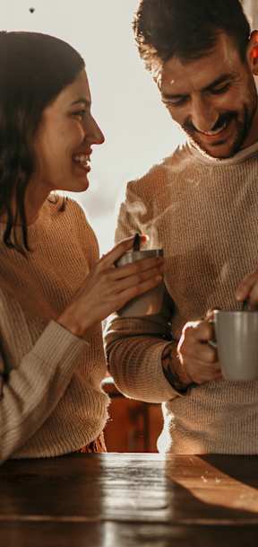 A man and a woman are standing at a table, with the man holding a mug and the woman holding a smartphone.