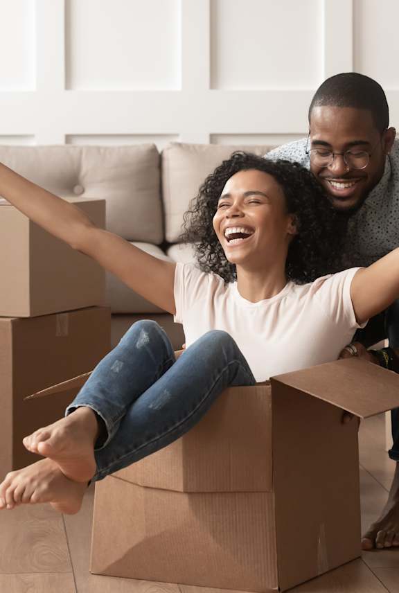 A man and woman are sitting on cardboard boxes in a room with a couch and plants.