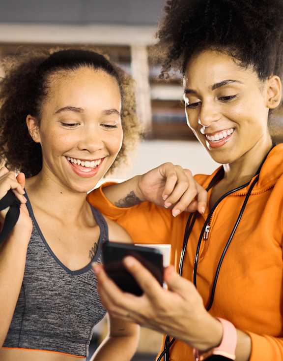 Two women wearing orange jackets and grey tops are looking at a smartphone.