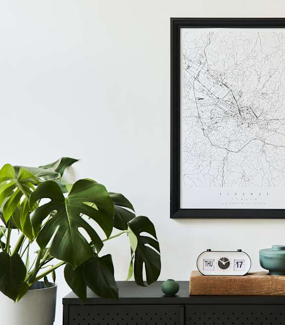 A black and white framed map is on the wall above a wooden table with a green pot, a clock, and a small potted plant.