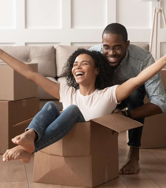A man and woman are sitting on cardboard boxes in a room with a plant and a lamp in the background.