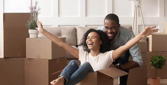 A man and woman are sitting on cardboard boxes in a room with a couch and plants.
