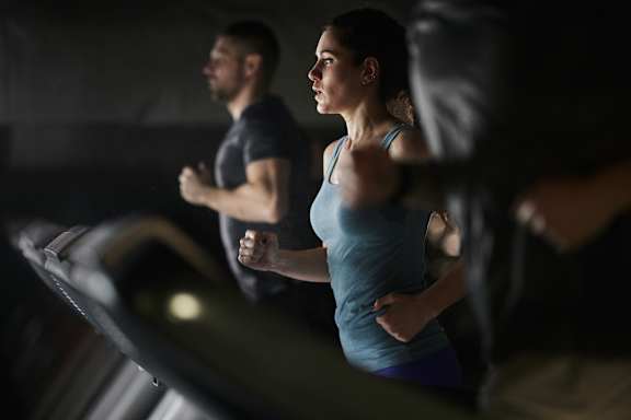 A woman in a blue tank top and purple leggings is running on a treadmill in a dark room.