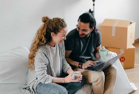 A man and a woman are sitting on a couch with a laptop, surrounded by cardboard boxes, suggesting they are in the process of moving in together.