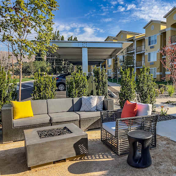The outdoor lounge with a table, plush chair, and couch next to a fire pit with the apartment buildings and covered parking lot at LIVEasy Street Apartments. 