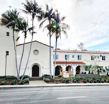 A white building with a red roof and palm trees in front.