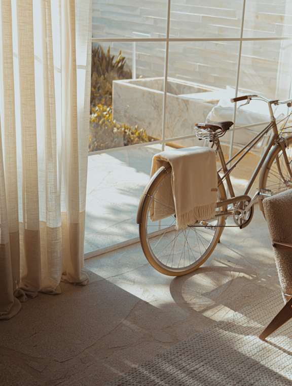 A bicycle with a towel hanging from the front basket is positioned in front of a window with sheer curtains.