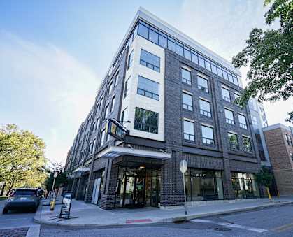 A modern multi-story building with a glass facade and a brick exterior is situated on a street corner with a car parked in front.