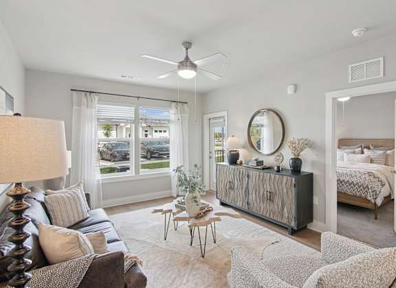 A well-lit, spacious living room with a large window, a sofa, a side table, and a bed in the background at The Hendry Apartments, Fort Myers, FL, 33905