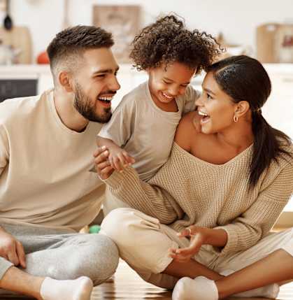 A man and a woman are sitting on the floor with a child between them, all are smiling and appear to be happy.