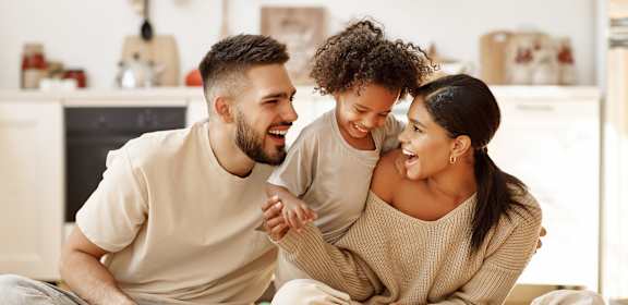 A man and a woman are sitting on the floor with a child between them, all are smiling and appear to be happy.