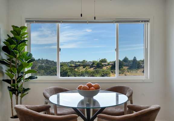 A table with a bowl of fruit on it sits in front of a window with a view of the outdoors.