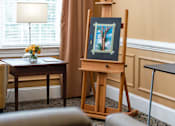 Thumbnail 3 of 5 - a painting on an easel in a living room with a couch and a table