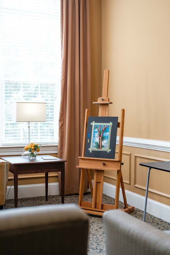 A painting of a tree is displayed on a wooden easel in a room with a table and a lamp.