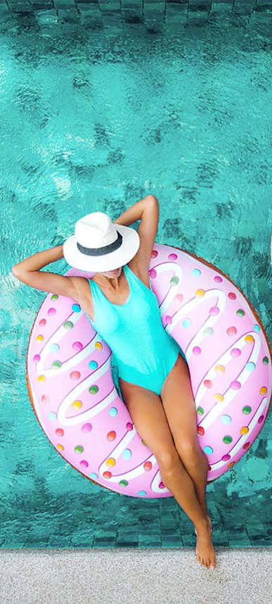 A woman in a blue swimsuit and white hat is floating on a pink donut-shaped float in a pool.